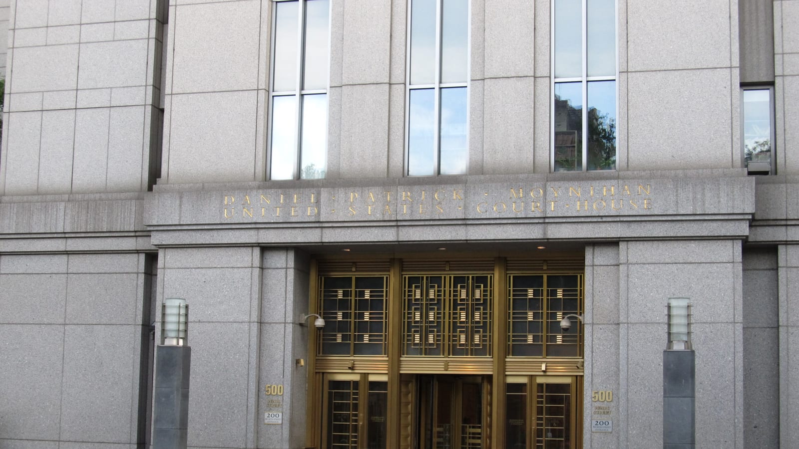 The gilded entrance doors of the Daniel Patrick Moynihan United States Courthouse under its carved name band, with the 500 Pearl Street number plates and bollards on the empty sidewalk.