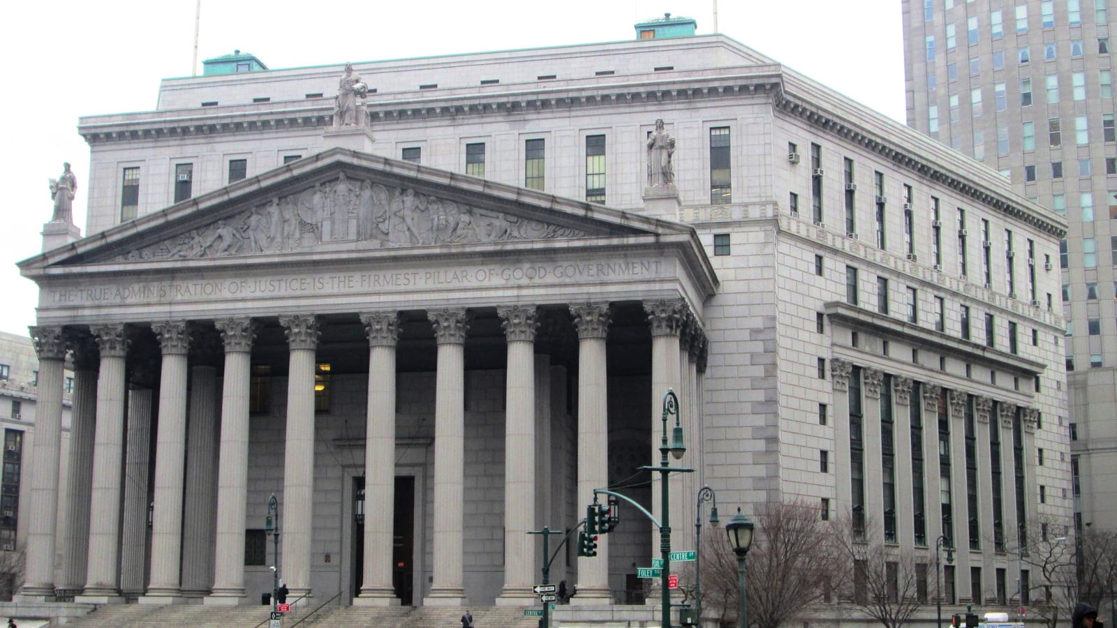 The Corinthian portico, carved pediment and wide stone steps of the New York County Courthouse at 60 Centre Street, photographed from the southwest on an overcast day with pedestrians crossing in front.