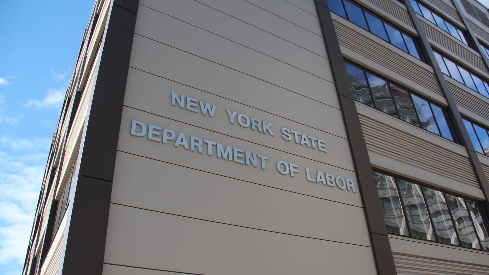 Metal lettering reading New York State Department of Labor across the upper facade of a tan panelled office building in Downtown Brooklyn, photographed from street level against a blue sky.