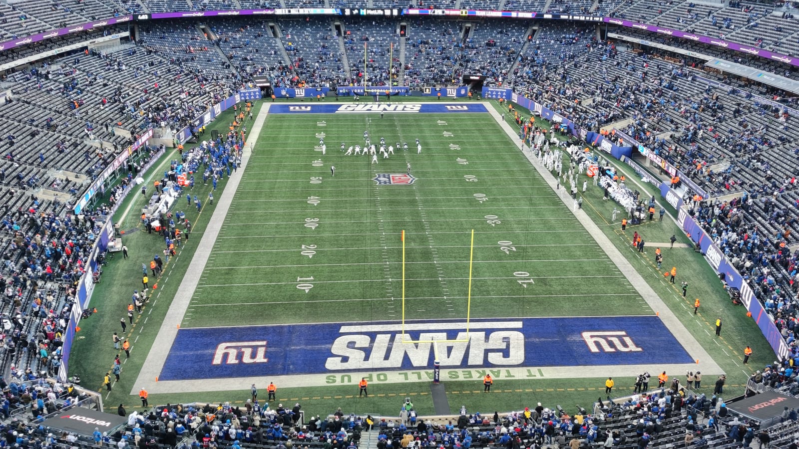 A wide view from the upper deck of a football stadium, looking down the length of the field toward an end zone painted blue with the word Giants, with players lined up near midfield and crowds in the stands.