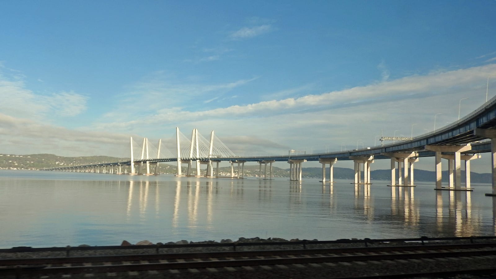 A long white cable-stayed bridge with paired towers crossing a wide, calm river under a blue sky, seen from beside railroad tracks on the near shore.