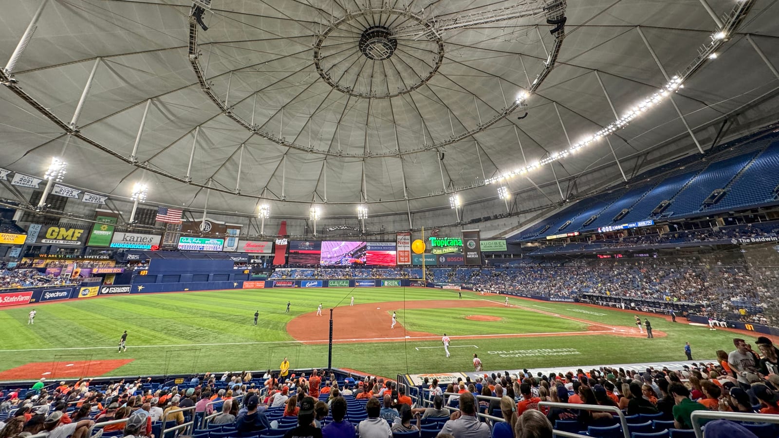 Interior of a domed ballpark during a game, with fans in the lower seats, players on the green field and a white fabric roof with a lit catwalk ring overhead.