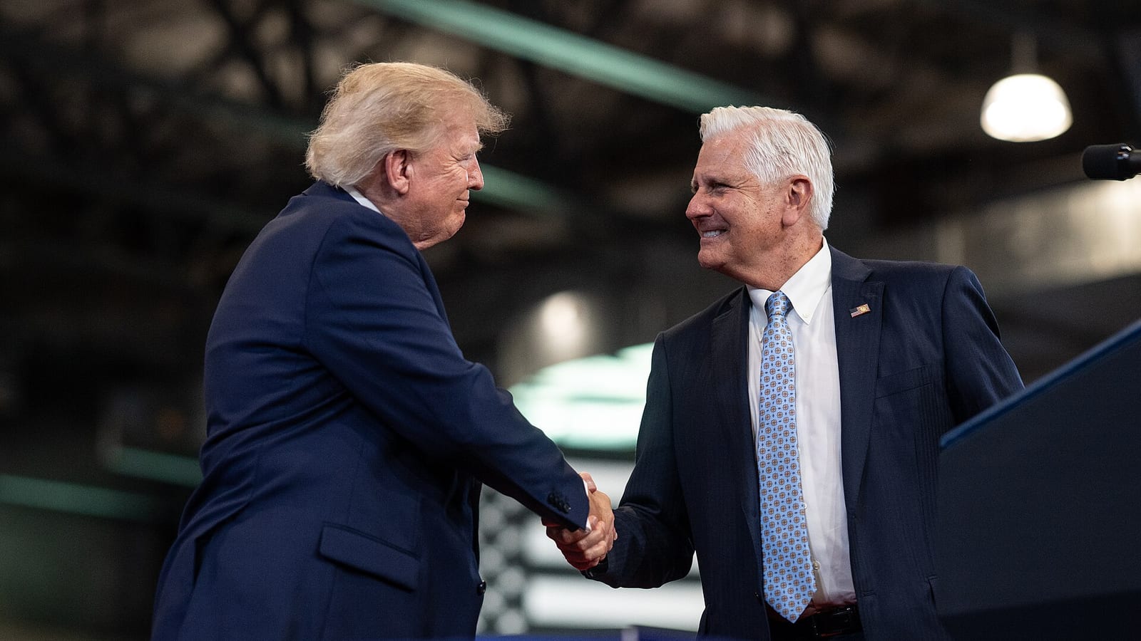 Two men in dark suits shake hands on a stage in front of a lectern, one with light blond hair on the left and one with white hair and a blue patterned tie on the right, above a blue banner.