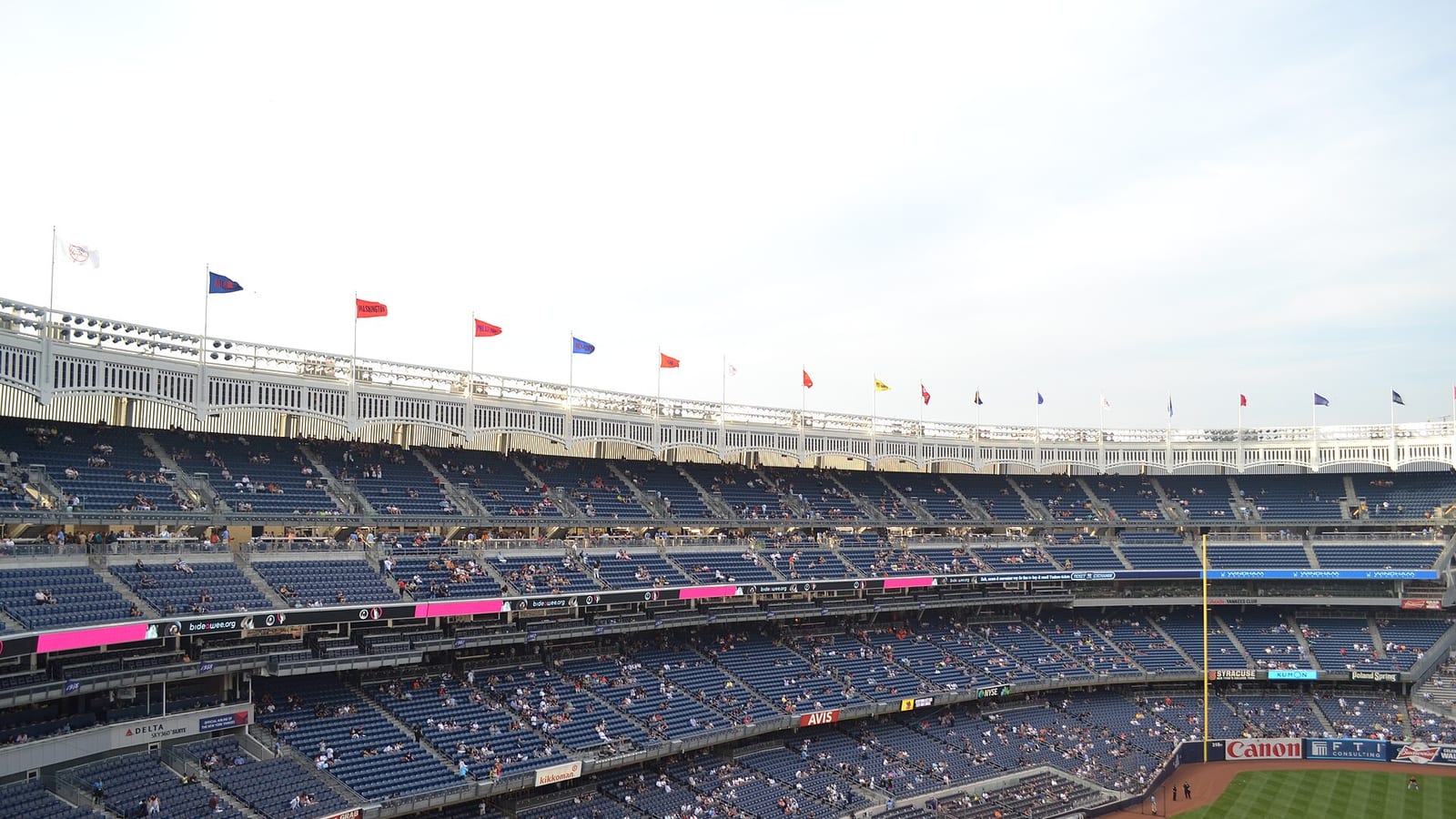 Tiers of mostly empty blue seats curving around a ballpark under a white decorative frieze lined with pennants, with the green outfield grass and a strip of infield dirt at the lower right.