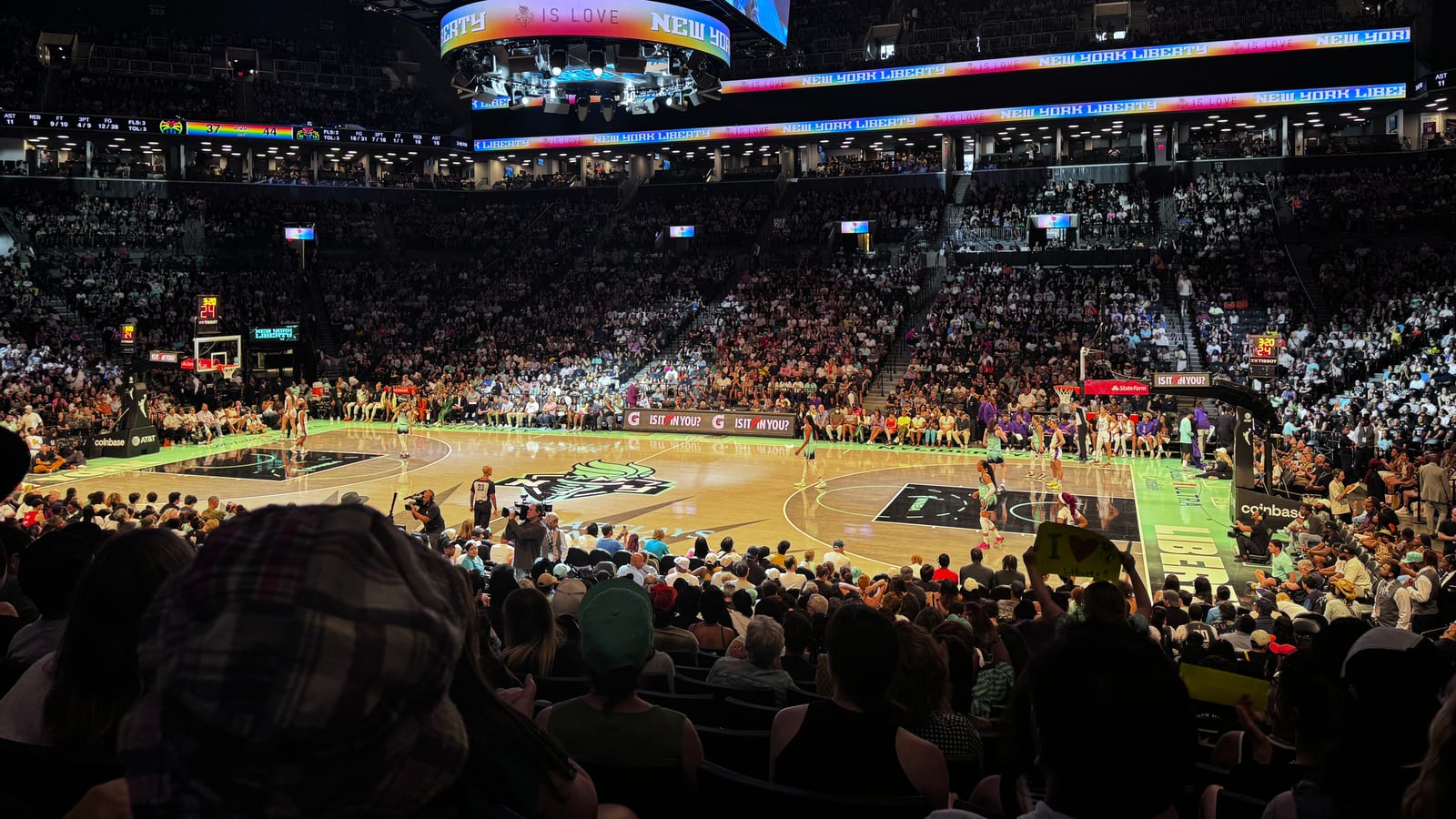A packed arena seen from courtside seats during a basketball game, with players on a court marked with the Liberty logo, a large video board overhead and ribbon displays reading New York Liberty around the upper bowl.