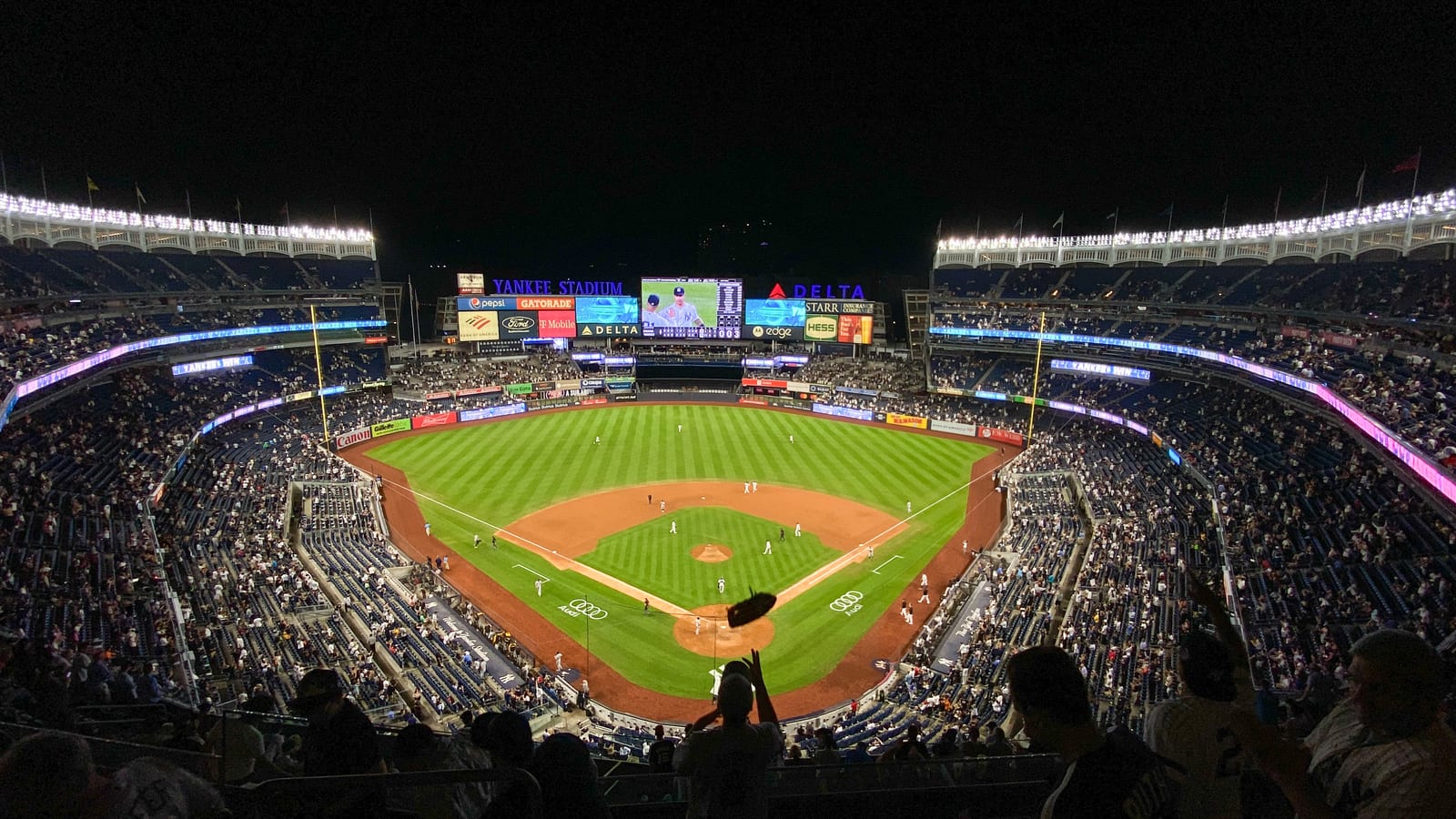 A night game seen from the upper deck behind home plate, with the lit infield and outfield grass, packed lower seating tiers and the Yankee Stadium scoreboard beyond center field.