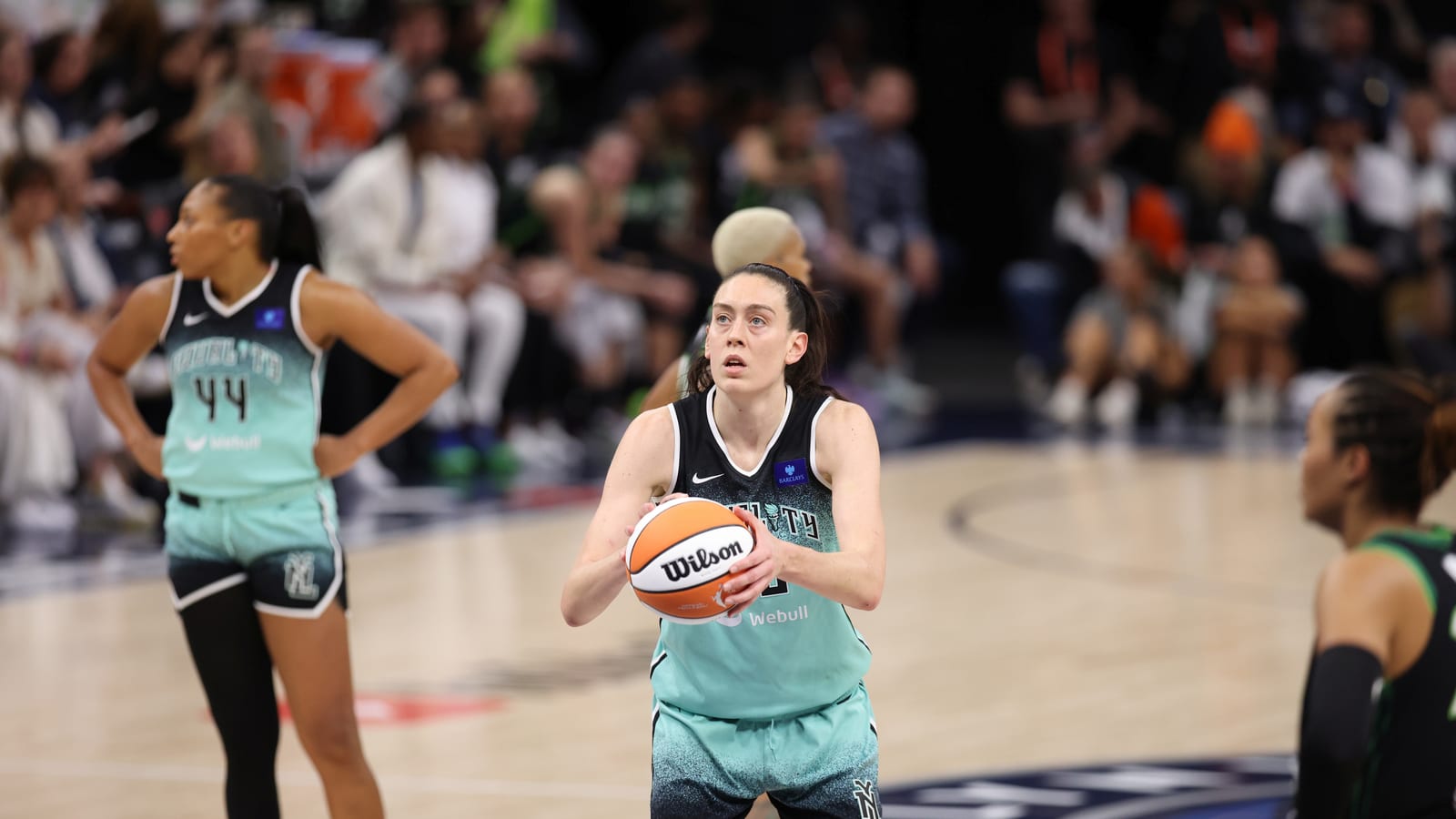 A basketball player in a mint green New York Liberty uniform holds the ball at the free-throw line, with a teammate standing behind her and an opponent in a dark uniform at the edge of the frame.