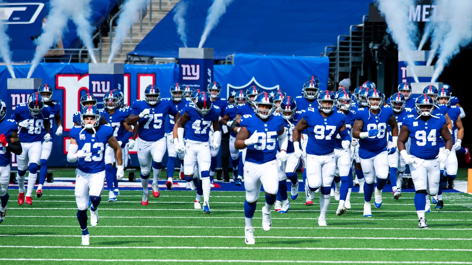 Football players in blue jerseys and white pants run onto a field through clouds of white smoke in front of blue banners bearing the NY logo.