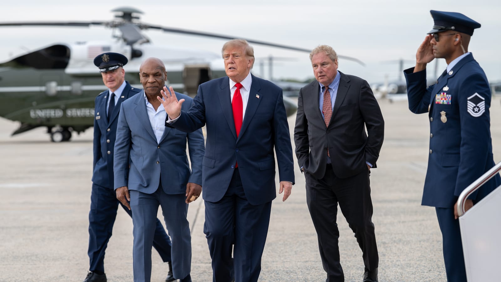 A man in a dark suit and red tie waves as he walks across an airfield tarmac with three other men, past a saluting member of the Air Force, with a green and white helicopter parked behind them.
