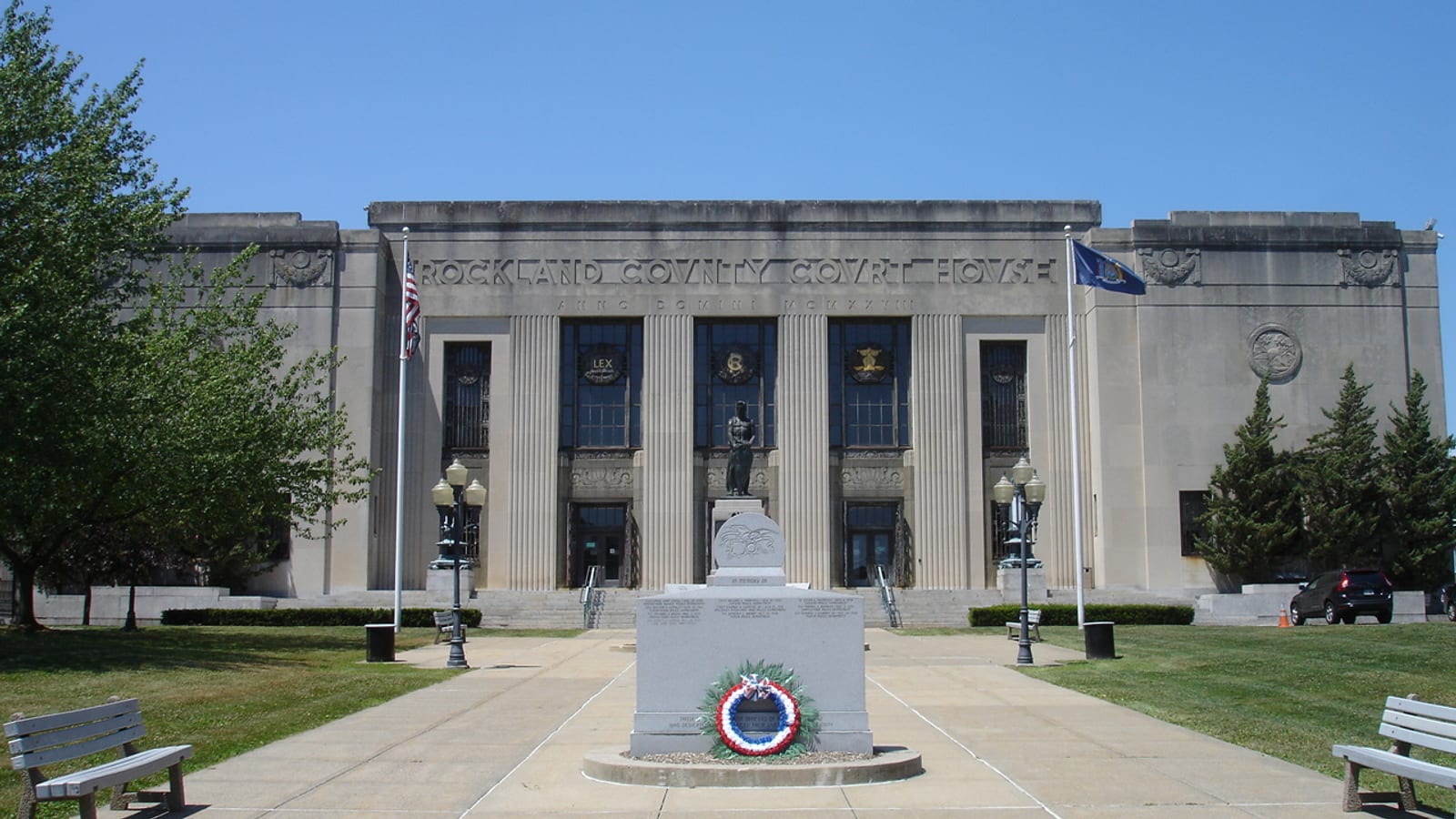 A pale stone courthouse with tall columns and the words Rockland County Court House carved above the entrance, with American and New York flags on either side and a war memorial with a red, white and blue wreath on the plaza in front.