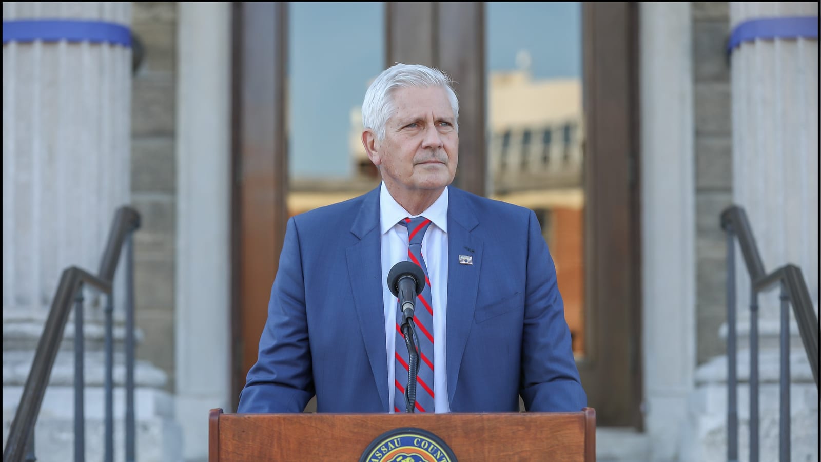 A man with white hair in a blue suit and striped red tie stands behind a wooden lectern bearing the Nassau County seal, in front of the columns and steps of a stone building.