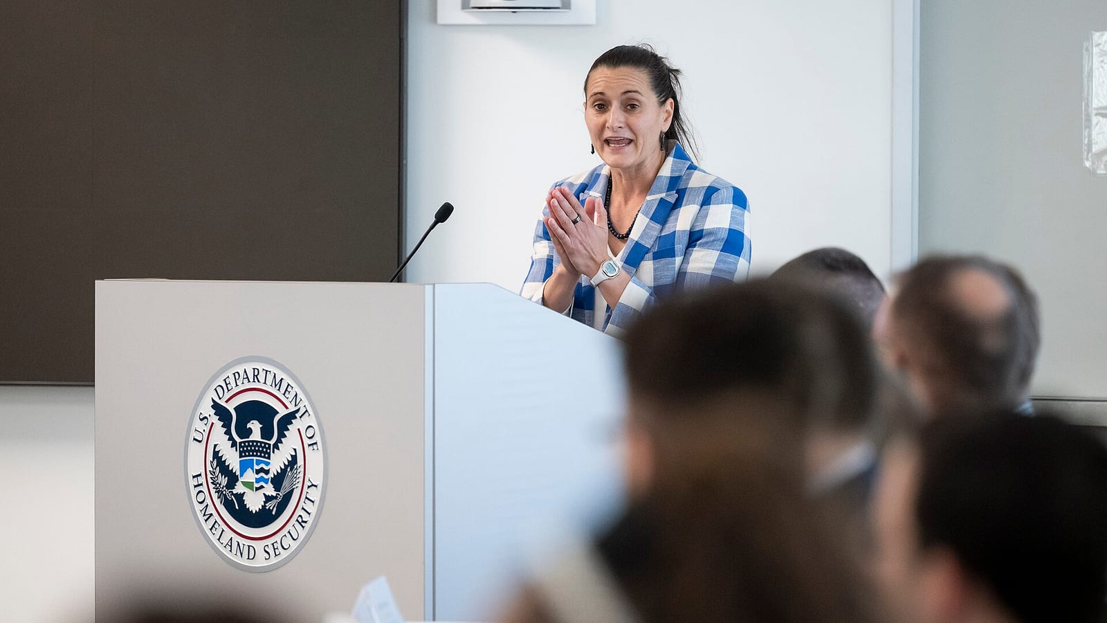A woman with dark hair tied back, wearing a blue and white checked jacket, speaks at a lectern bearing the U.S. Department of Homeland Security seal, with audience members blurred in the foreground.